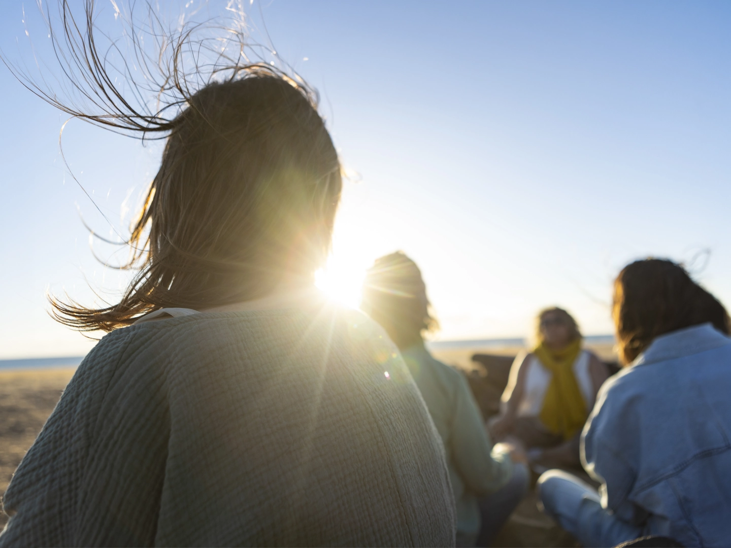 Grupo de personas reunidas en una playa al atardecer. En primer plano aparece una persona de espaldas, con el cabello movido por el viento y el sol brillando entre los mechones. Al fondo, varias personas están sentadas en círculo cerca de la orilla, iluminadas por la luz cálida del sol bajo sobre el horizonte.