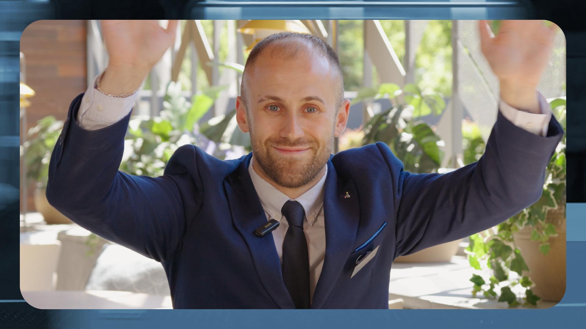 Persona con traje azul marino y corbata posa frente a la cámara con ambos brazos levantados. Se encuentra en un espacio interior luminoso con plantas y grandes ventanales al fondo. La imagen está enmarcada dentro de un diseño gráfico con bordes redondeados y tonos azules.