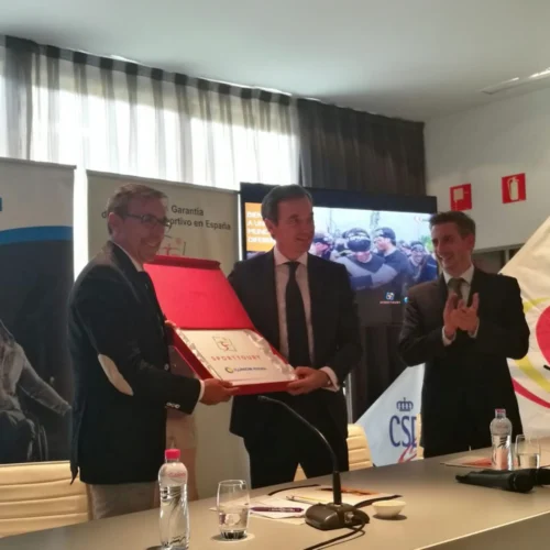 Tres personas posan durante un acto institucional en una sala de conferencias. Dos de ellas sostienen una placa o reconocimiento enmarcado, mientras la tercera aplaude. En primer plano hay una mesa con micrófonos, vasos y botellas de agua, y al fondo se observan paneles informativos, una pantalla y una vela con logotipos relacionados con el deporte y la inclusión.