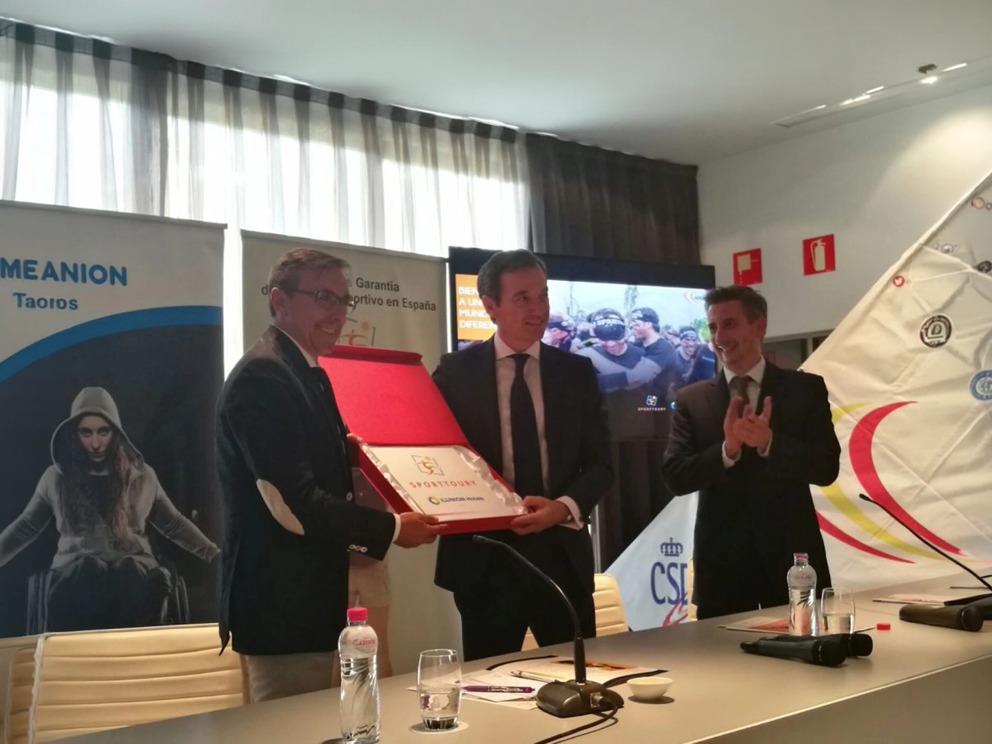 Tres personas posan durante un acto institucional en una sala de conferencias. Dos de ellas sostienen una placa o reconocimiento enmarcado, mientras la tercera aplaude. En primer plano hay una mesa con micrófonos, vasos y botellas de agua, y al fondo se observan paneles informativos, una pantalla y una vela con logotipos relacionados con el deporte y la inclusión.
