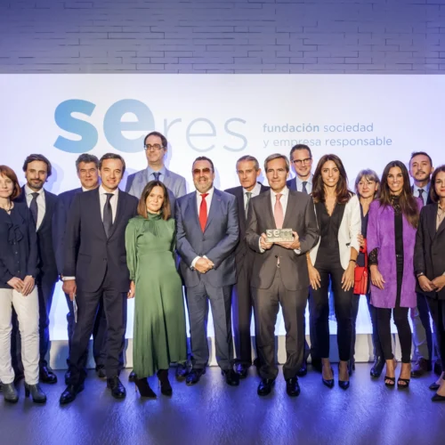 Grupo de personas posando en un escenario durante un acto institucional, delante de un panel con el logotipo de “SERES, fundación sociedad y empresa responsable”. En el centro, una persona sostiene un reconocimiento mientras el resto del grupo se distribuye alrededor. La escena está iluminada con luces azules y blancas.