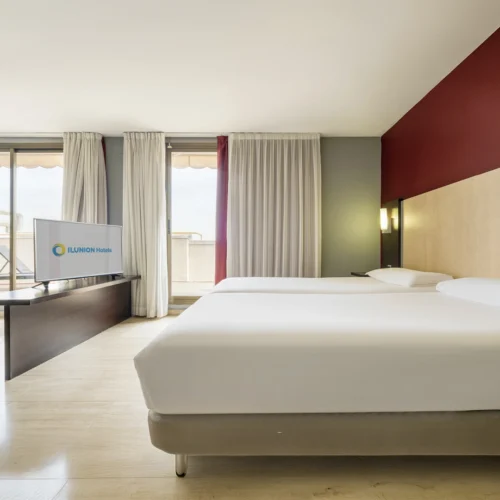 Habitación de hotel amplia y luminosa con dos camas individuales de sábanas blancas. Frente a las camas hay un televisor con el logotipo de ILUNION Hotels sobre un mueble oscuro. Grandes ventanales con cortinas blancas dan acceso a una terraza exterior, mientras una pared en tono burdeos y un cabecero de madera clara completan la decoración moderna del espacio.