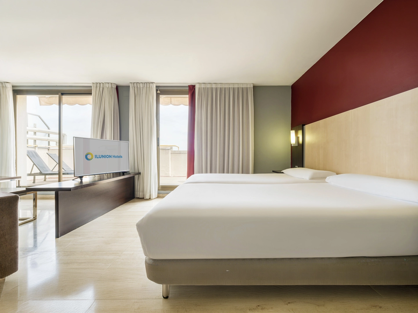 Habitación de hotel amplia y luminosa con dos camas individuales de sábanas blancas. Frente a las camas hay un televisor con el logotipo de ILUNION Hotels sobre un mueble oscuro. Grandes ventanales con cortinas blancas dan acceso a una terraza exterior, mientras una pared en tono burdeos y un cabecero de madera clara completan la decoración moderna del espacio.