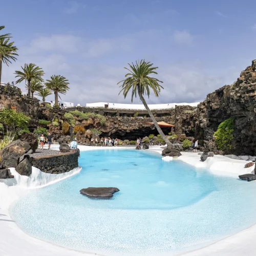 Lagunilla de agua turquesa diseñada en forma orgánica, rodeada de roca volcánica negra, palmeras altas y senderos de piedra; varias personas pasean o descansan en los bordes dentro de un espacio abierto y luminoso.