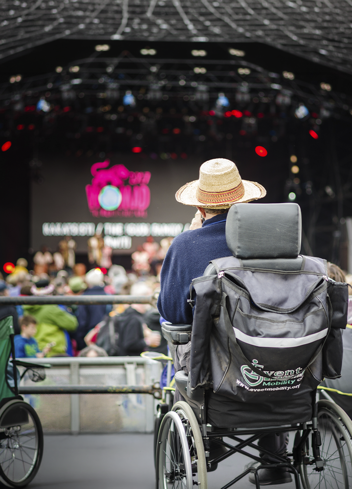 Persona en silla de ruedas observa un concierto o evento multitudinario desde una zona reservada frente al escenario. Lleva un sombrero de paja y ocupa el primer plano de la imagen, mientras al fondo se distinguen el público, las luces del recinto y una gran pantalla desenfocada. La escena transmite un entorno accesible e inclusivo en un festival al aire libre.