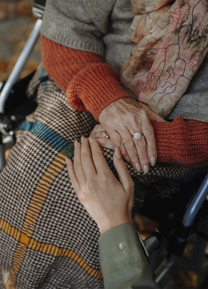 Primer plano de una persona sentada en una silla de ruedas con una manta de cuadros sobre las piernas y las manos apoyadas sobre el regazo. Otra persona acerca una mano en gesto de acompañamiento y apoyo. La escena destaca el contacto entre ambas manos, mientras se aprecian prendas de abrigo en tonos cálidos y un entorno exterior desenfocado al fondo.
