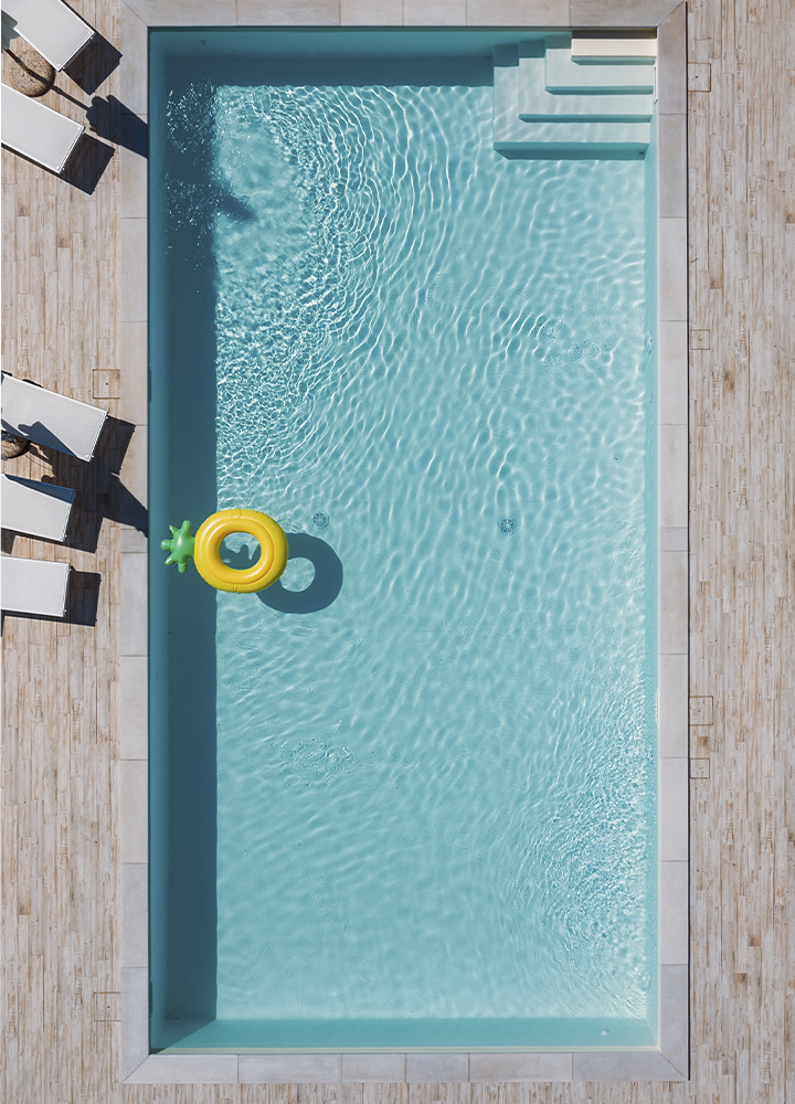Vista cenital de una piscina rectangular de agua cristalina, rodeada por un solárium de madera clara. En una esquina flotan un hinchable amarillo con forma de piña y su sombra proyectada sobre el fondo de la piscina. Varias tumbonas blancas se alinean a ambos lados, mientras unos escalones integrados permiten el acceso al agua en el extremo superior. La luz del sol crea ondulaciones y reflejos sobre toda la superficie azul.