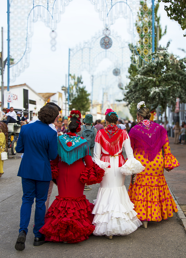 Personas caminan por una avenida decorada para una feria o celebración popular. En primer plano aparecen varias de espaldas, vestidas con trajes tradicionales de colores vivos y mantones bordados con flecos. Destacan un vestido rojo, otro blanco y otro amarillo con lunares rosas, acompañados de flores en el cabello. La calle está llena de asistentes y se encuentra adornada con arcos luminosos, árboles y casetas a ambos lados, bajo un cielo claro durante el día.
