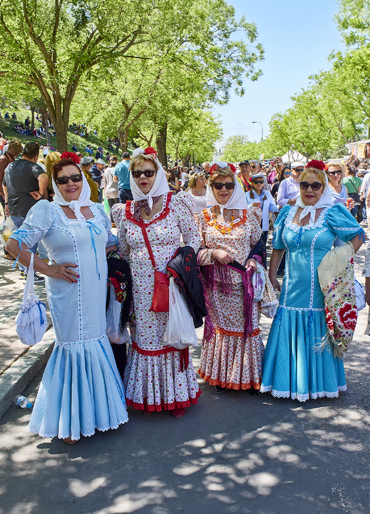 Cuatro personas posan en una avenida arbolada durante una fiesta popular al aire libre. Llevan vestidos tradicionales de colores claros y estampados florales, acompañados de pañuelos blancos en la cabeza, adornos florales rojos y gafas de sol. Alrededor se observa una gran afluencia de público paseando por el recinto, con árboles frondosos que proporcionan sombra y puestos o actividades al fondo.