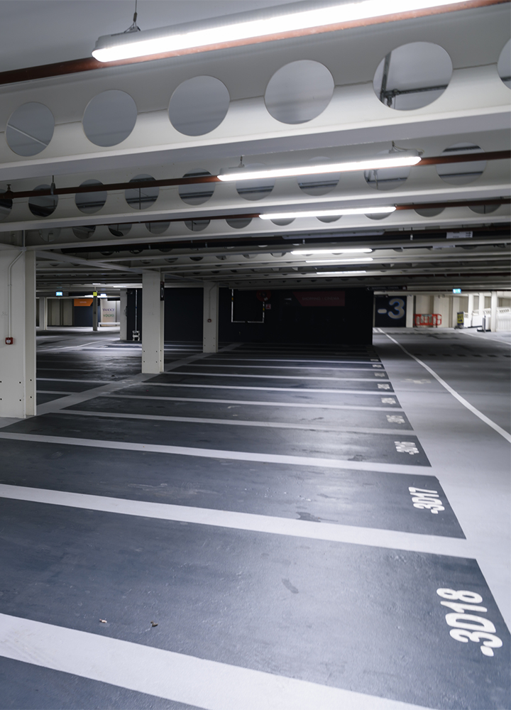 Aparcamiento subterráneo prácticamente vacío, con largas filas de plazas delimitadas por líneas blancas sobre el suelo oscuro. En el lateral derecho se distinguen los números de las plazas pintados en blanco y, al fondo, un gran indicador con el número “-3” señala la planta del estacionamiento. El espacio está iluminado por filas de luces fluorescentes en el techo y sostenido por columnas blancas, creando una perspectiva profunda y ordenada.