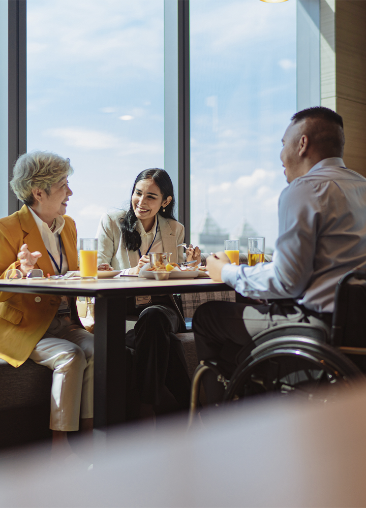 Tres personas comparten una comida o reunión en una mesa junto a grandes ventanales con vistas al exterior. Una de ellas utiliza una silla de ruedas. Sobre la mesa hay vasos con zumo, platos y utensilios, mientras las credenciales colgadas al cuello sugieren un entorno profesional o de evento. La luz natural ilumina el espacio y crea un ambiente abierto y acogedor.