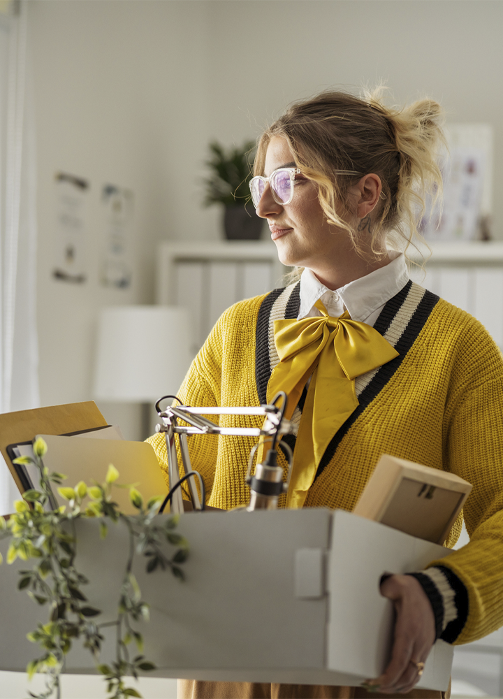 Persona con gafas sostiene una caja con documentos, una lámpara de escritorio y otros objetos de oficina en un espacio de trabajo luminoso. Viste un cárdigan amarillo sobre una camisa blanca con un lazo del mismo color y se encuentra junto a una ventana que deja entrar luz natural. Al fondo se observan estanterías con archivadores, plantas decorativas y mobiliario de oficina en tonos claros.