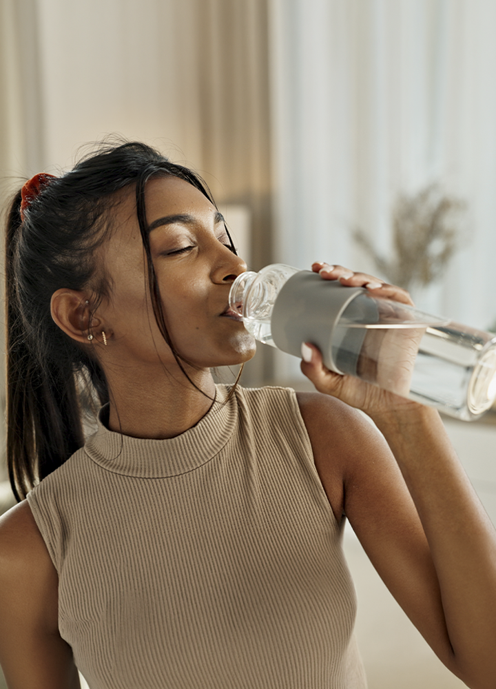 Primer plano de una persona bebiendo agua de una botella reutilizable transparente con funda de silicona gris. La botella está inclinada hacia la boca y contiene agua visible en su interior. La persona viste una camiseta sin mangas de canalé en color beige y lleva el cabello recogido. El fondo aparece desenfocado, con cortinas y elementos decorativos en tonos neutros, mientras la luz cálida del interior resalta la botella y la acción de hidratarse.
