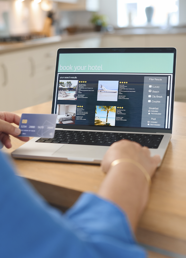 Persona utilizando un ordenador portátil para realizar una reserva de hotel mientras sostiene una tarjeta bancaria en una mano. En la pantalla aparece una página de búsqueda de alojamientos con el texto “book your hotel”, fotografías de hoteles, valoraciones con estrellas y filtros de búsqueda relacionados con tipo de viaje, desayuno y piscina. El portátil está sobre una mesa de madera en un entorno doméstico luminoso, con una cocina desenfocada al fondo. La escena transmite una gestión de reservas o compras de viajes por internet.