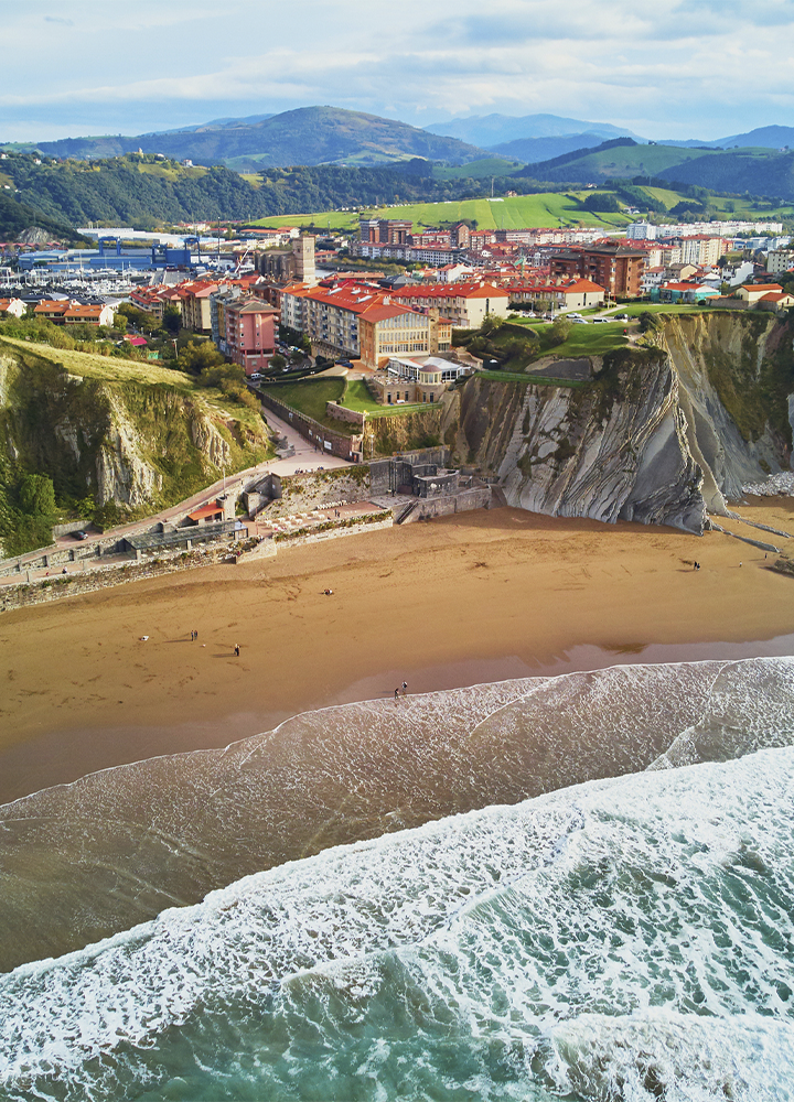 Vista aérea de una playa rodeada por espectaculares acantilados de roca estratificada junto a una localidad costera. Las olas rompen sobre la arena mientras un paseo marítimo recorre la base de los acantilados. Al fondo se extiende el núcleo urbano con edificios de tejados rojizos, un puerto y colinas verdes que se elevan hasta las montañas del horizonte bajo un cielo parcialmente nublado.