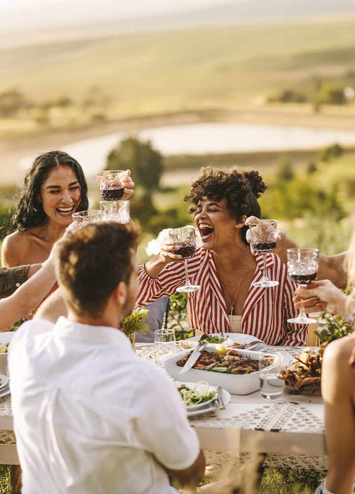 Grupo de personas reunidas alrededor de una mesa al aire libre brindando con copas de vino tinto. La mesa está preparada con distintos platos de comida y bebida, mientras al fondo se extiende un paisaje natural con zonas verdes y un lago desenfocado. La escena está iluminada por la luz cálida del atardecer, que aporta tonos dorados y un ambiente de celebración y encuentro.