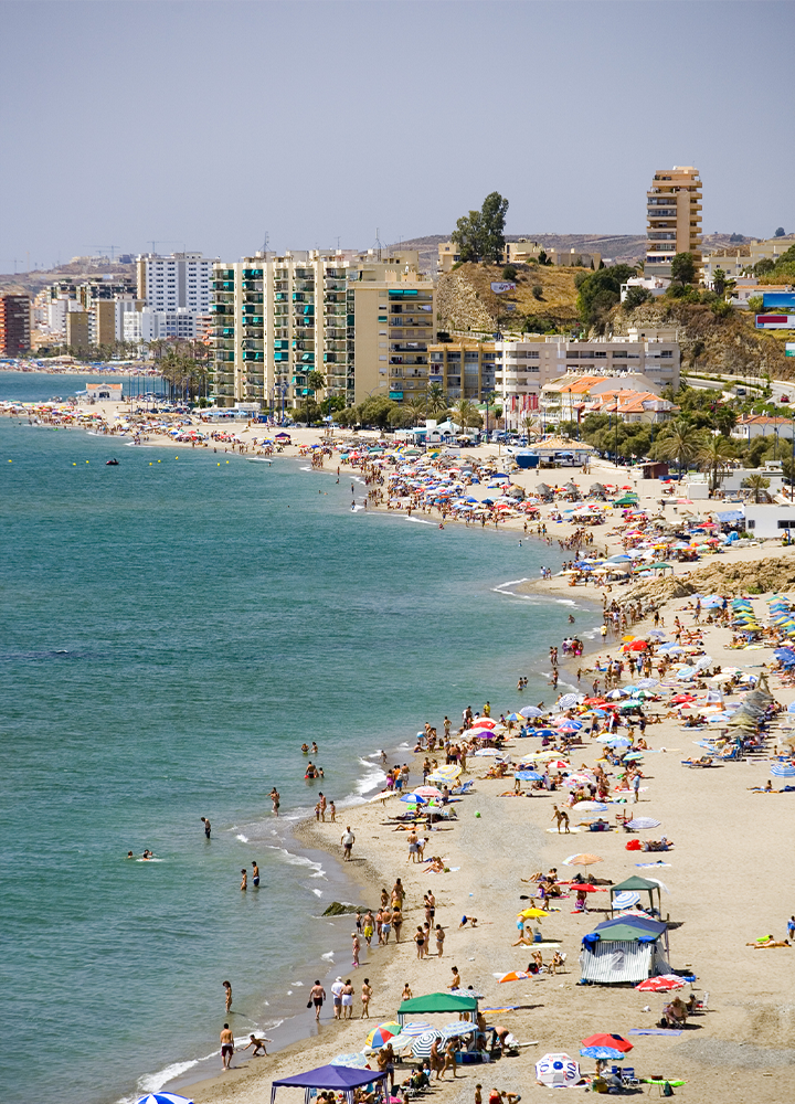 Playa urbana muy concurrida con numerosas personas distribuidas a lo largo de la orilla y del agua. La costa forma una amplia curva junto al mar en calma, mientras sombrillas de colores y pequeñas carpas se reparten por la arena. Al fondo se alzan edificios residenciales y hoteles de varias plantas, algunos situados sobre una ladera con vegetación. La escena está iluminada por luz intensa de día bajo un cielo despejado.