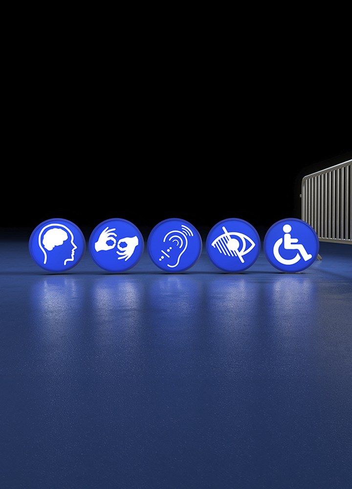 Cinco iconos azules de accesibilidad alineados sobre una superficie brillante. Representan accesibilidad cognitiva, lengua de signos, accesibilidad auditiva, accesibilidad visual y movilidad reducida. Al fondo se observa una valla metálica y un espacio oscuro que resalta los símbolos, cuyos reflejos se proyectan sobre el suelo iluminado en tonos azules.