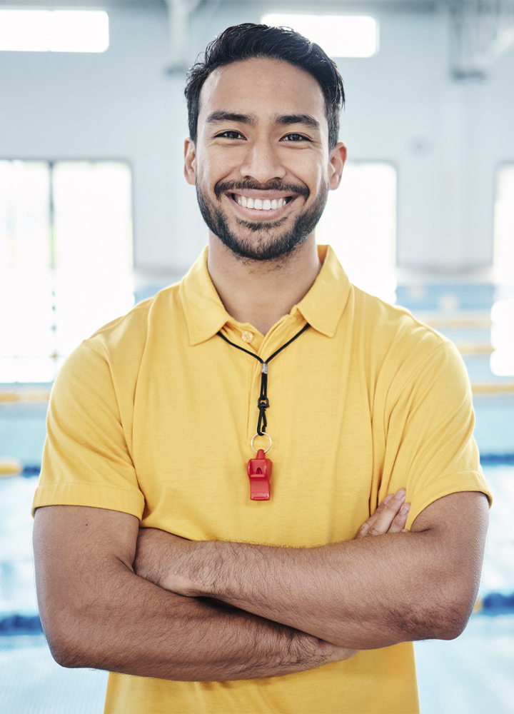 Persona con los brazos cruzados posa frente a una piscina cubierta. Lleva un polo amarillo y un silbato rojo colgado al cuello. El fondo aparece desenfocado, con grandes ventanales y las líneas de las calles de nado visibles, creando un entorno deportivo e interior iluminado por luz natural.