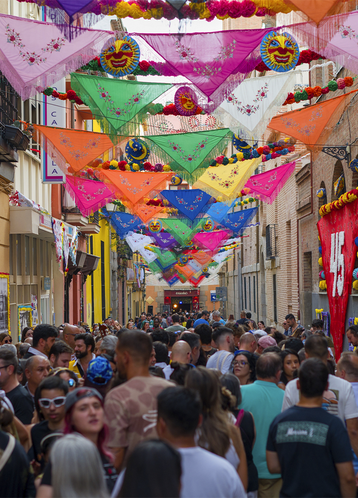 Calle estrecha repleta de personas durante una celebración popular. Sobre la vía cuelgan numerosos mantones y adornos de colores vivos, en tonos rosa, verde, naranja, azul, amarillo y blanco, decorando el espacio de un lado a otro entre los edificios. Entre los adornos destacan figuras circulares con diseños de sol y guirnaldas de flores de colores. A ambos lados se alinean fachadas de comercios y viviendas, mientras la multitud llena prácticamente toda la calle.