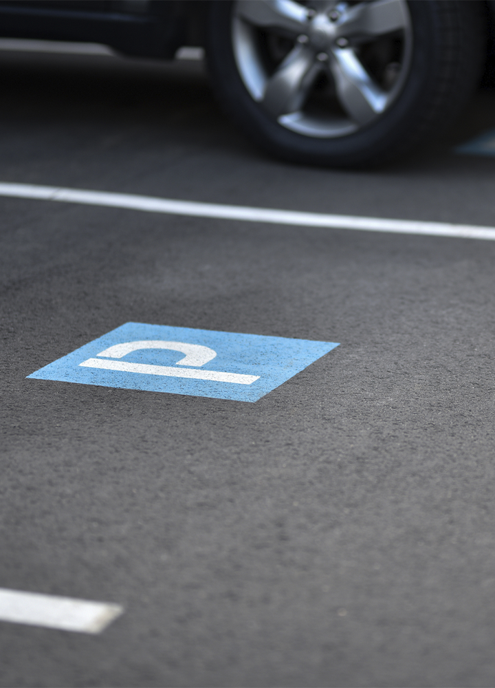 Plaza de aparcamiento reservada para personas con movilidad reducida, señalizada con el símbolo de accesibilidad pintado en blanco sobre un fondo azul en el asfalto. En la parte superior de la imagen se aprecia parcialmente la rueda de un vehículo estacionado, mientras las líneas blancas delimitan las plazas sobre la superficie gris del aparcamiento.