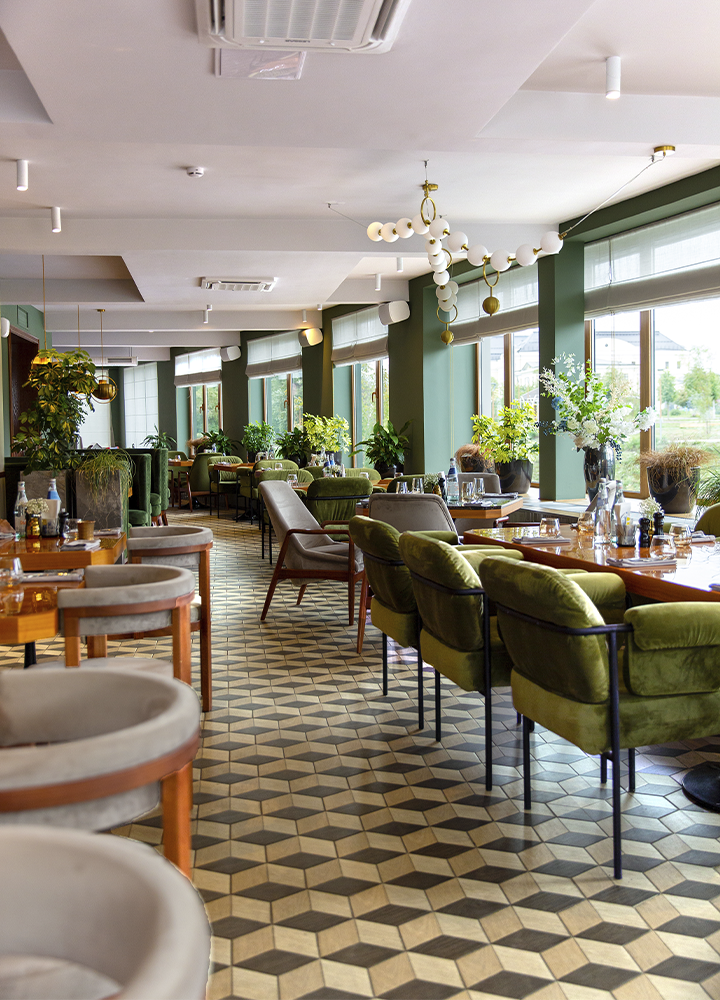 Restaurante amplio y luminoso con decoración contemporánea en tonos verdes y madera. Varias mesas están preparadas para el servicio, rodeadas de sillones tapizados en verde y sillas de diseño claro. Grandes ventanales recorren uno de los laterales, dejando entrar abundante luz natural y ofreciendo vistas a una zona ajardinada. El espacio se completa con numerosas plantas, lámparas decorativas suspendidas del techo y un suelo geométrico en tonos beige y gris que aporta profundidad y elegancia al conjunto.