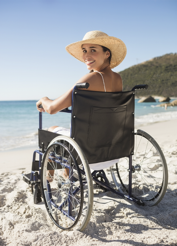 Persona sentada en una silla de ruedas sobre la arena de una playa, junto a la orilla del mar. Lleva un sombrero de ala ancha y ropa clara, y gira la cabeza hacia la cámara mientras sostiene una de las ruedas de la silla. Al fondo se observan el agua, una costa rocosa y una ladera cubierta de vegetación bajo un cielo despejado y luminoso. La escena transmite accesibilidad y disfrute de un entorno costero al aire libre.