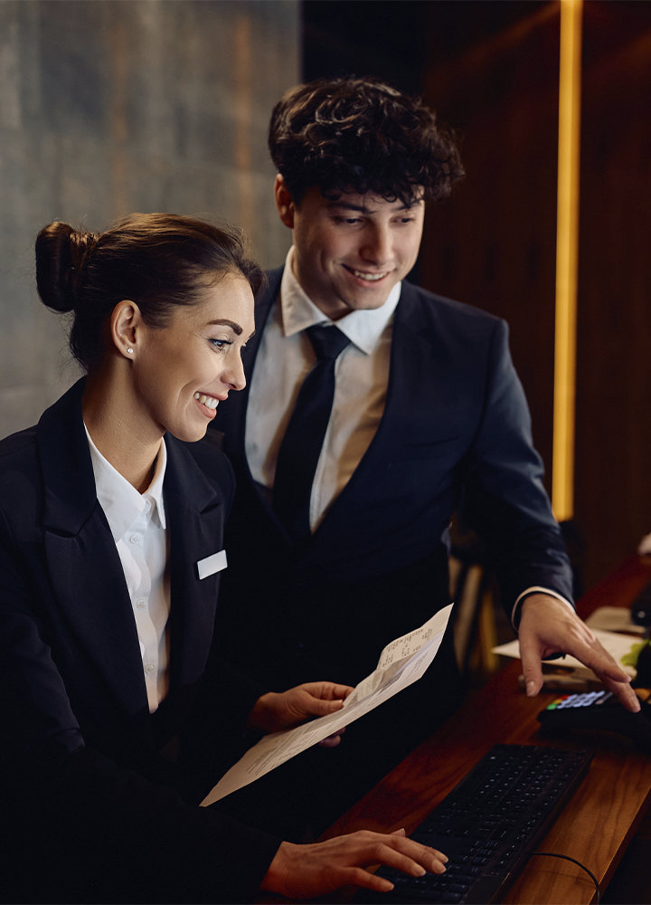 Dos personas con indumentaria formal trabajando detrás de un mostrador de recepción. Una de ellas sostiene varios documentos mientras utiliza un teclado, y la otra señala una pantalla o terminal situado fuera de la imagen. El entorno presenta iluminación cálida, mobiliario de madera y acabados elegantes propios de un hotel o espacio de atención al cliente. La interacción se centra en la gestión de información o el proceso de registro, con equipos informáticos y documentación visibles sobre el mostrador.