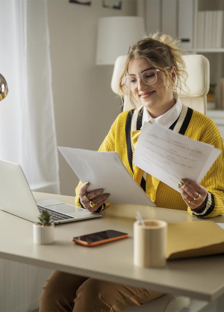 Persona sentada en un despacho luminoso revisa varios documentos mientras trabaja frente a un ordenador portátil. Lleva gafas y un jersey amarillo sobre una camisa blanca con un lazo del mismo color. Sobre la mesa hay un teléfono móvil, una libreta, un pequeño tiesto y algunos papeles. Al fondo se observan estanterías con archivadores y una lámpara de pie en un entorno de oficina ordenado y tranquilo.