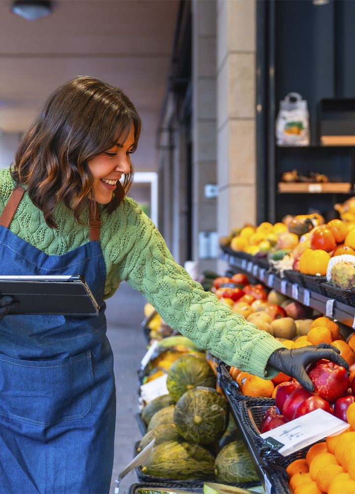 Persona con delantal azul y guantes oscuros seleccionando una manzana roja en la sección de frutas y verduras de una tienda. En una mano sostiene una tableta y, frente a ella, hay estantes repletos de manzanas, naranjas, melones y otros productos frescos organizados en cajas. El establecimiento está iluminado con luz cálida y el fondo aparece desenfocado, destacando la actividad de revisión o reposición de la fruta.