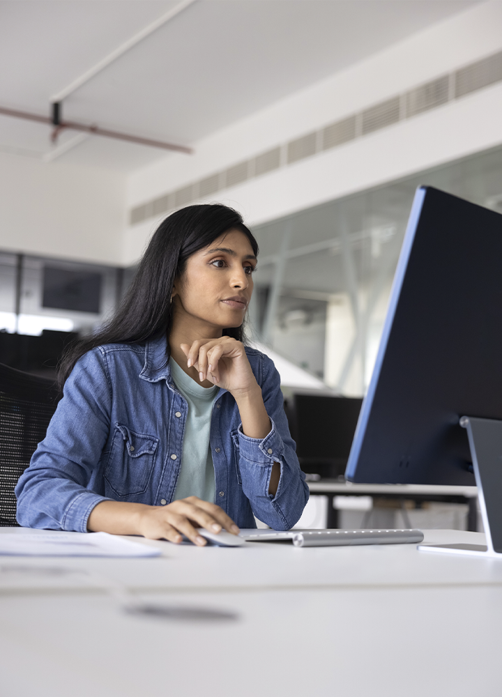 Persona sentada frente a un monitor de gran tamaño en una oficina moderna. Una mano descansa sobre un ratón y la otra sostiene la barbilla mientras observa la pantalla. Sobre el escritorio blanco se aprecia un teclado y algunos documentos desenfocados en primer plano. El espacio cuenta con paredes acristaladas y mobiliario de oficina en tonos neutros, iluminado por abundante luz natural que aporta un ambiente de trabajo profesional y despejado.