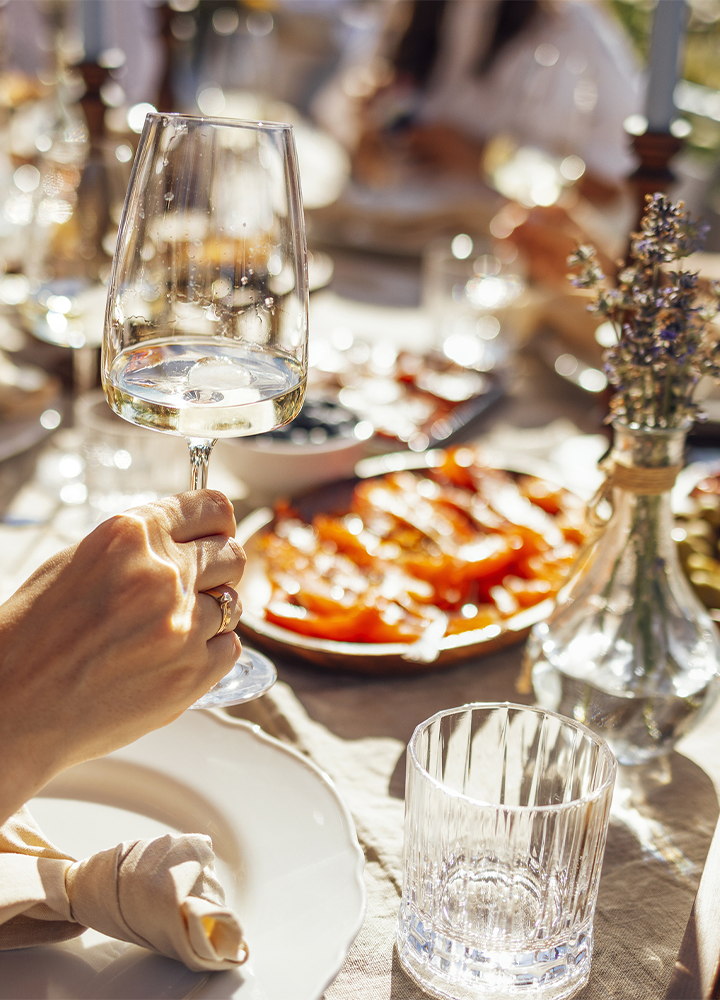 Detalle de una mesa preparada para una comida al aire libre. En primer plano, una mano sostiene una copa de vino blanco sobre un mantel claro con vajilla, cristalería y una servilleta enrollada. Al fondo aparecen platos con comida desenfocados y un pequeño jarrón con flores silvestres, iluminados por una luz cálida que crea reflejos y destellos sobre el vidrio.