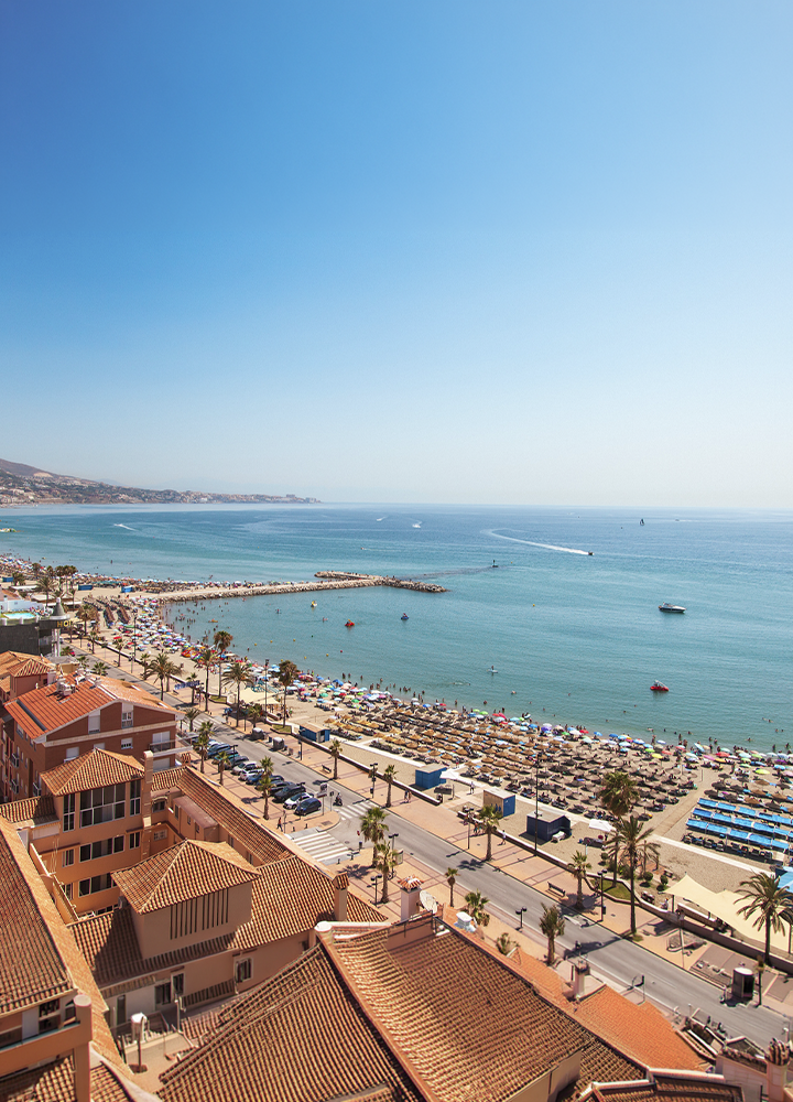 Vista elevada de una playa urbana junto a una amplia bahía de aguas tranquilas. En primer plano destacan edificios de tejados rojizos y un paseo marítimo bordeado de palmeras. La playa está llena de sombrillas y bañistas, y un espigón de piedra se adentra en el mar formando una pequeña zona resguardada. Al fondo se extiende la costa con colinas suaves y varias embarcaciones navegando sobre el agua bajo un cielo despejado.