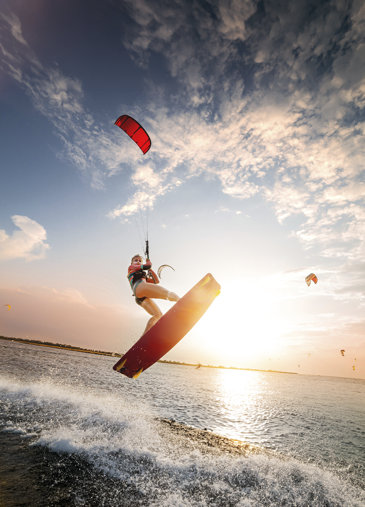 Persona practicando kitesurf realiza un salto sobre el agua al atardecer. Sujeta la cometa mediante un arnés mientras la tabla se eleva por encima de las olas, con salpicaduras en primer plano. El sol bajo ilumina la escena desde el horizonte y varias cometas se distinguen a lo lejos bajo un cielo amplio con nubes dispersas.
