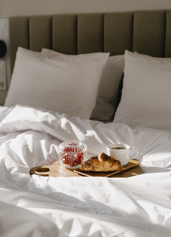 Cama con ropa de cama blanca y almohadas grandes apoyadas sobre un cabecero acolchado oscuro. En el centro de la cama hay una bandeja de madera con una taza de café, un cruasán y un recipiente de cristal con fruta roja. La luz natural ilumina las sábanas arrugadas y crea sombras suaves, transmitiendo una escena de desayuno servido en una habitación de hotel o alojamiento.