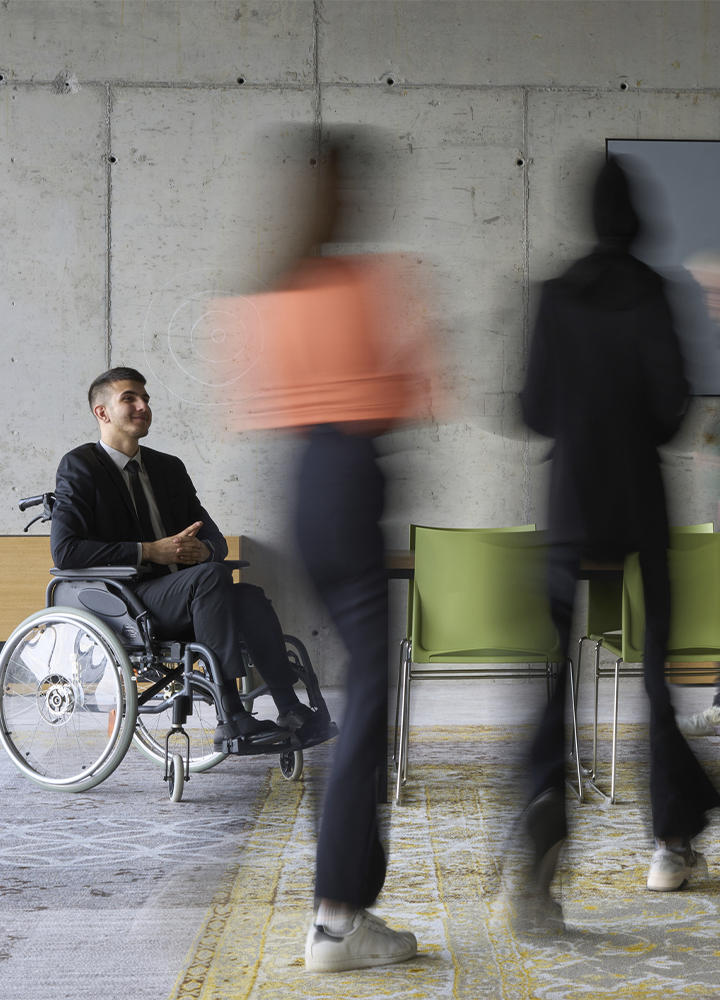 Persona en silla de ruedas espera en una sala de reuniones de estilo moderno junto a una mesa y varias sillas verdes. En primer plano, varias personas pasan caminando y aparecen desenfocadas por el movimiento, creando contraste con la persona sentada, que permanece nítida. El espacio cuenta con una pared de hormigón visto, suelo con alfombra de tonos grises y amarillos, y mobiliario contemporáneo.