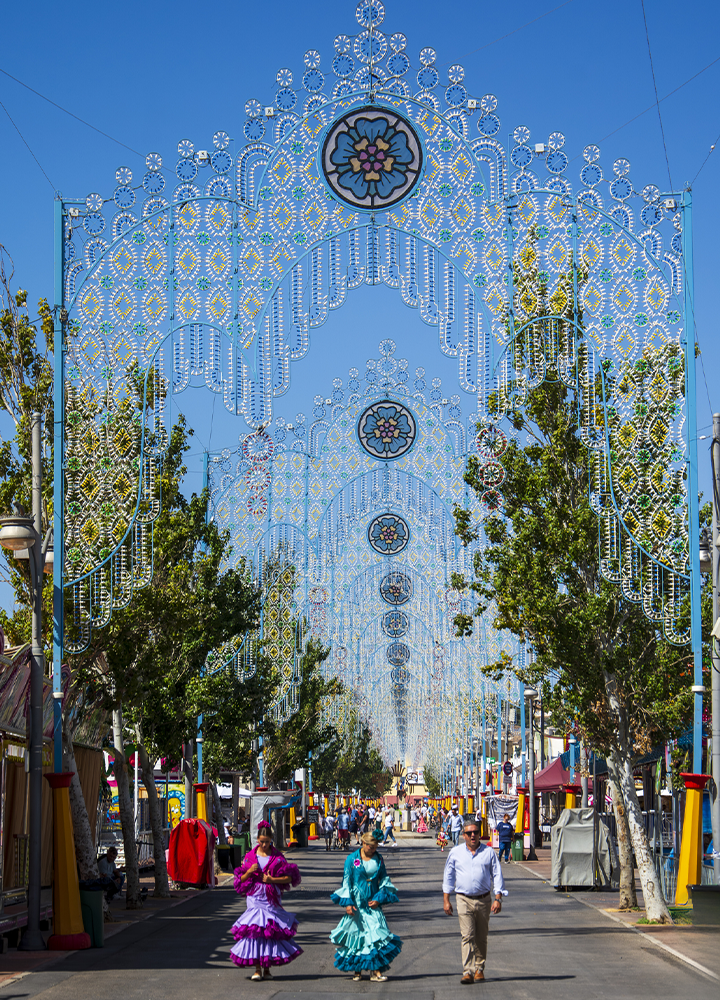 Avenida amplia decorada con grandes arcos de luces ornamentales que se repiten a lo largo de la calle bajo un cielo azul despejado. A ambos lados hay árboles, casetas y estructuras festivas. En primer plano, varias personas caminan por el centro de la avenida, algunas con trajes tradicionales de volantes en colores vivos. La perspectiva de los arcos crea un efecto de profundidad que conduce la vista hacia el fondo, donde continúa la actividad de la celebración popular.