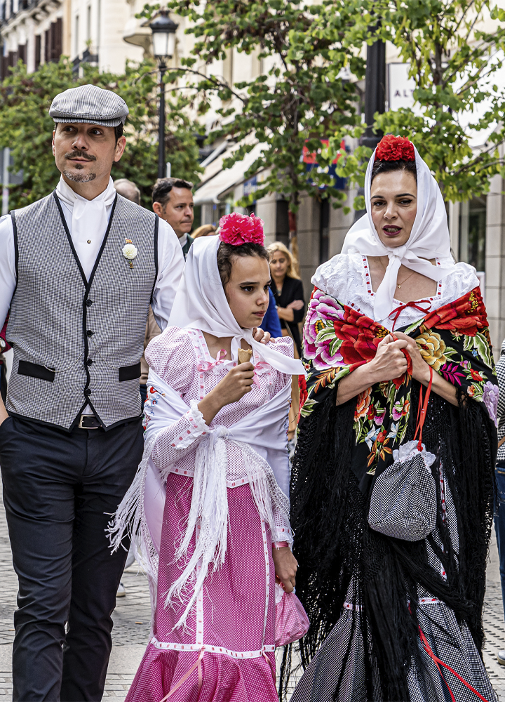 Tres personas caminan por una calle peatonal durante una celebración tradicional. Visten trajes regionales con mantones bordados, pañuelos blancos sobre la cabeza y adornos florales de colores. La persona situada en el centro sostiene un helado, mientras las otras dos caminan a ambos lados. Al fondo se observan árboles, farolas, escaparates y varias personas paseando por la calle.
