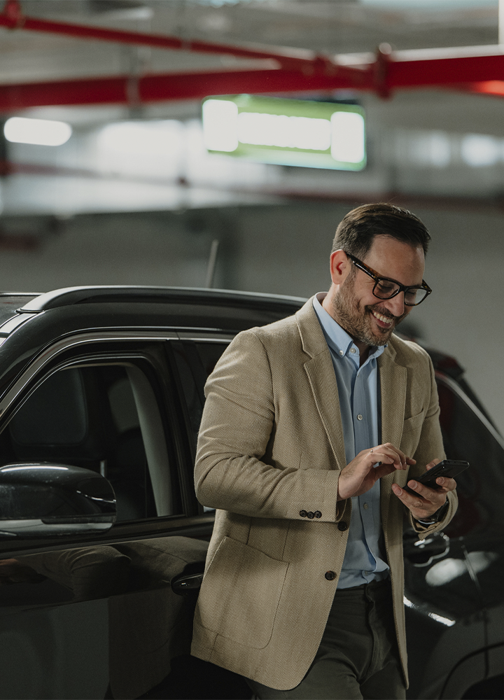 Persona apoyada junto a un vehículo en un aparcamiento cubierto mientras utiliza un teléfono móvil. Viste americana beige, camisa azul clara y gafas. El coche oscuro ocupa gran parte del lateral izquierdo de la imagen, mientras el fondo aparece desenfocado con iluminación artificial, señales luminosas y estructuras rojas en el techo. La escena transmite un entorno de movilidad y uso de tecnología en un estacionamiento interior.