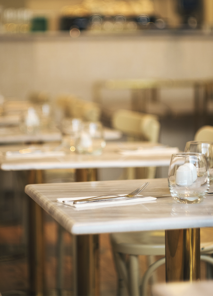Detalle de varias mesas preparadas en un restaurante, con manteles claros, vasos de cristal y cubiertos colocados sobre servilletas blancas. En primer plano destaca una mesa de mármol con un tenedor y dos vasos, mientras el resto del comedor aparece suavemente desenfocado al fondo. La iluminación cálida y los tonos beige crean un ambiente elegante y tranquilo.