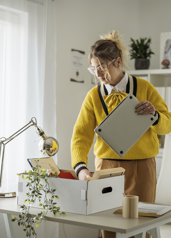 Persona con gafas recoge objetos de una caja de cartón sobre una mesa mientras sostiene un ordenador portátil cerrado. Viste un jersey amarillo sobre una camisa blanca y se encuentra en un espacio de trabajo luminoso con una lámpara de escritorio, una planta colgante y estanterías al fondo. La escena transmite un momento de recogida o traslado de pertenencias en una oficina.