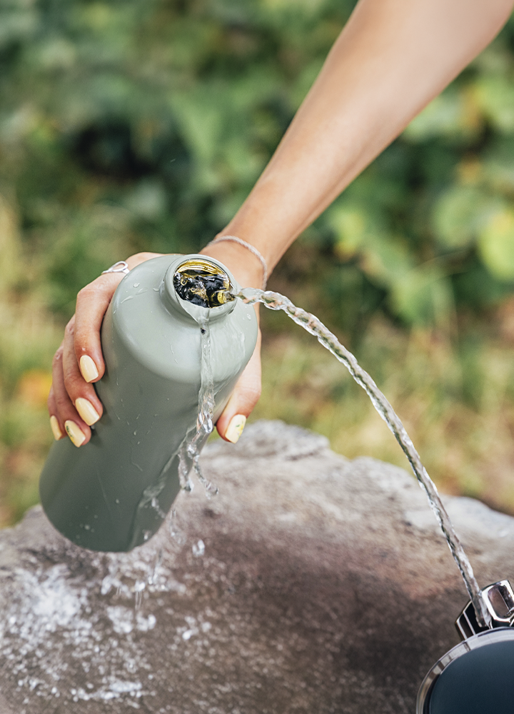Primer plano de una mano sujetando una botella reutilizable gris mientras vierte agua en un entorno exterior. El agua sale por la abertura de la botella formando dos corrientes que caen sobre una superficie de piedra. La mano lleva una pulsera fina y las uñas están pintadas en tono claro. El fondo aparece desenfocado, con vegetación verde que aporta profundidad y sitúa la escena en un espacio natural al aire libre.