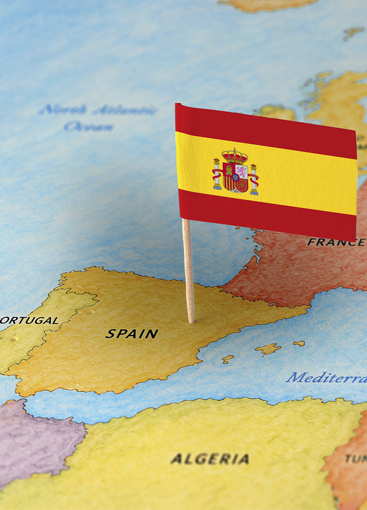 Bandera de España clavada sobre un mapa de la península ibérica, con el mástil situado en territorio español. Se distinguen los nombres de España, Portugal, Francia y Argelia, además del mar Mediterráneo y el océano Atlántico Norte. La bandera, de franjas roja, amarilla y roja con el escudo nacional en el centro, ocupa el primer plano sobre un mapa de colores suaves.