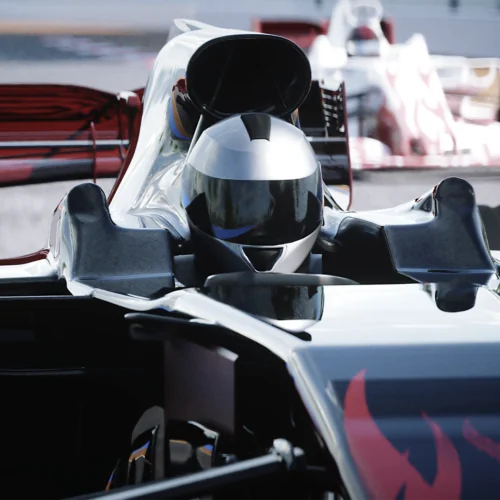 Primer plano frontal de un monoplaza de competición en un circuito de carreras. El vehículo presenta una carrocería aerodinámica en colores negro, plata y rojo, con un casco de piloto visible en el habitáculo. Destacan el alerón delantero, las tomas de aire superiores y los neumáticos anchos característicos de este tipo de vehículos. Al fondo aparece otro monoplaza desenfocado, reforzando el contexto de competición automovilística. La iluminación resalta las superficies brillantes y las líneas técnicas del vehículo, que ocupa casi toda la imagen.