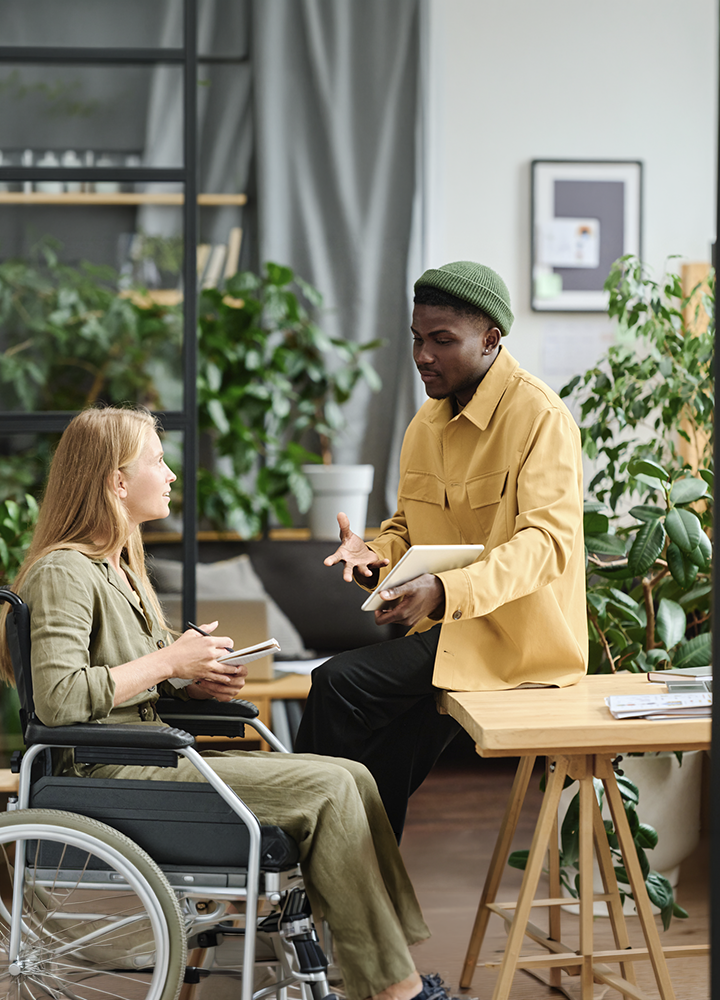 Dos personas conversan en una oficina rodeada de plantas. Una está sentada en una silla de ruedas tomando notas en una libreta, mientras la otra se apoya en una mesa de madera y sostiene una tableta digital. El espacio es luminoso, con estanterías, cortinas grises y abundante vegetación que aporta un ambiente moderno y accesible.
