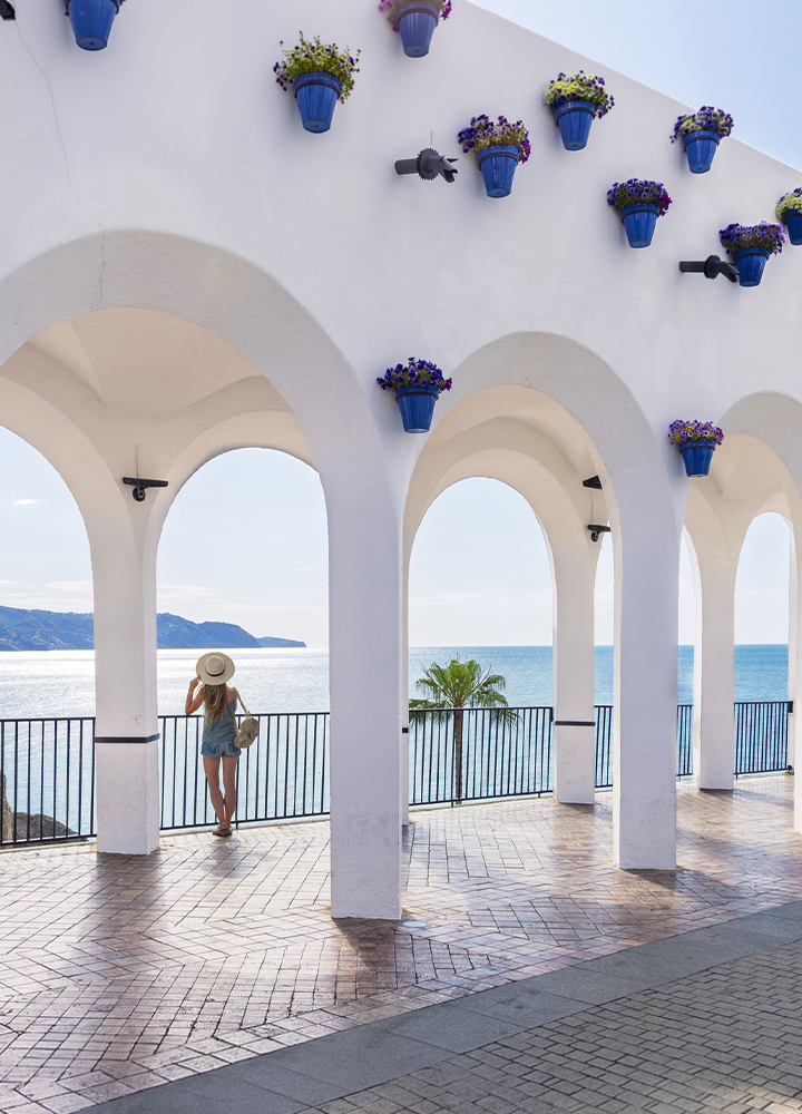 Persona con sombrero contempla el mar desde un mirador cubierto por arcos blancos de estilo mediterráneo. Las paredes están decoradas con macetas azules llenas de flores, mientras una barandilla negra separa la terraza del agua. Al fondo se distinguen la costa, una pequeña isla y una palmera, bajo una luz suave y un cielo despejado.
