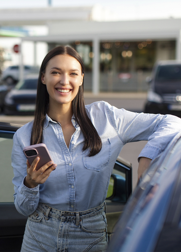 Persona apoyada en la puerta de un coche mientras sostiene un teléfono móvil. Viste una camisa vaquera clara y unos vaqueros de cintura alta. El vehículo aparece parcialmente en primer plano y, al fondo, se observan otros coches aparcados y un edificio desenfocados. La escena está iluminada por luz natural y tiene un aire de movilidad y uso de tecnología en un entorno urbano.