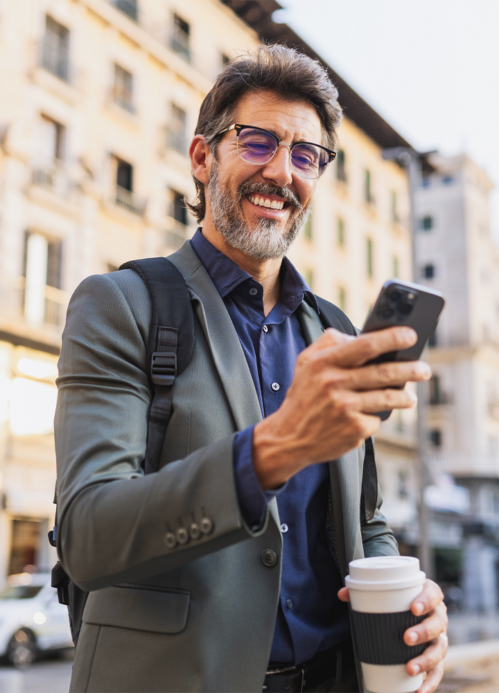 Persona con gafas, chaqueta y mochila utilizando un teléfono móvil mientras sostiene un vaso de café para llevar. La escena transcurre en una calle urbana con edificios desenfocados al fondo y luz natural cálida. El teléfono ocupa el centro de la acción, mientras la mochila y el café sugieren un desplazamiento o actividad cotidiana en la ciudad.