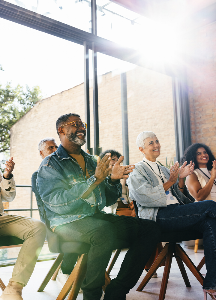 Grupo de personas sentadas en sillas durante una presentación o evento en un espacio interior con grandes ventanales. Varias personas aplauden mientras miran hacia un punto situado fuera de la imagen. La sala cuenta con abundante luz natural que entra por los ventanales y crea destellos en la parte superior. Al fondo se observan paredes de ladrillo visto y estructuras metálicas, aportando un estilo moderno e industrial. La composición se centra en la audiencia y en la interacción colectiva durante la actividad.