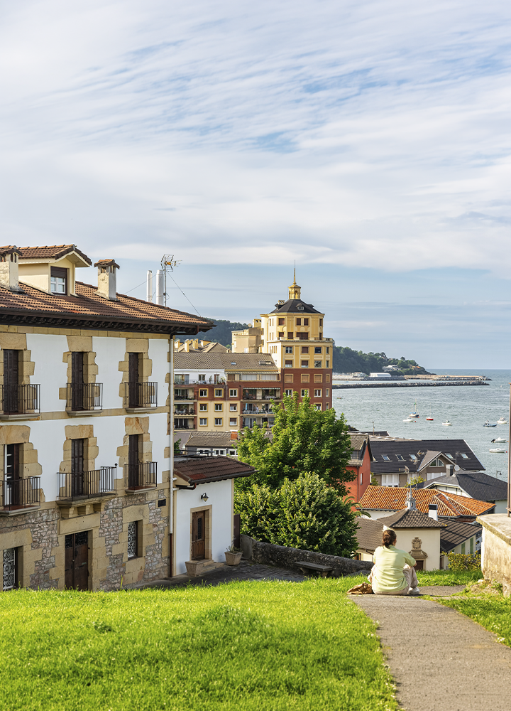 Vista elevada de una localidad costera con casas de tejados rojizos y edificios junto al mar. En primer plano, una persona sentada sobre un camino contempla la bahía desde una zona ajardinada. Al fondo se observan el puerto, varias embarcaciones sobre el agua y una línea de costa iluminada por la luz del día bajo un cielo parcialmente nublado.